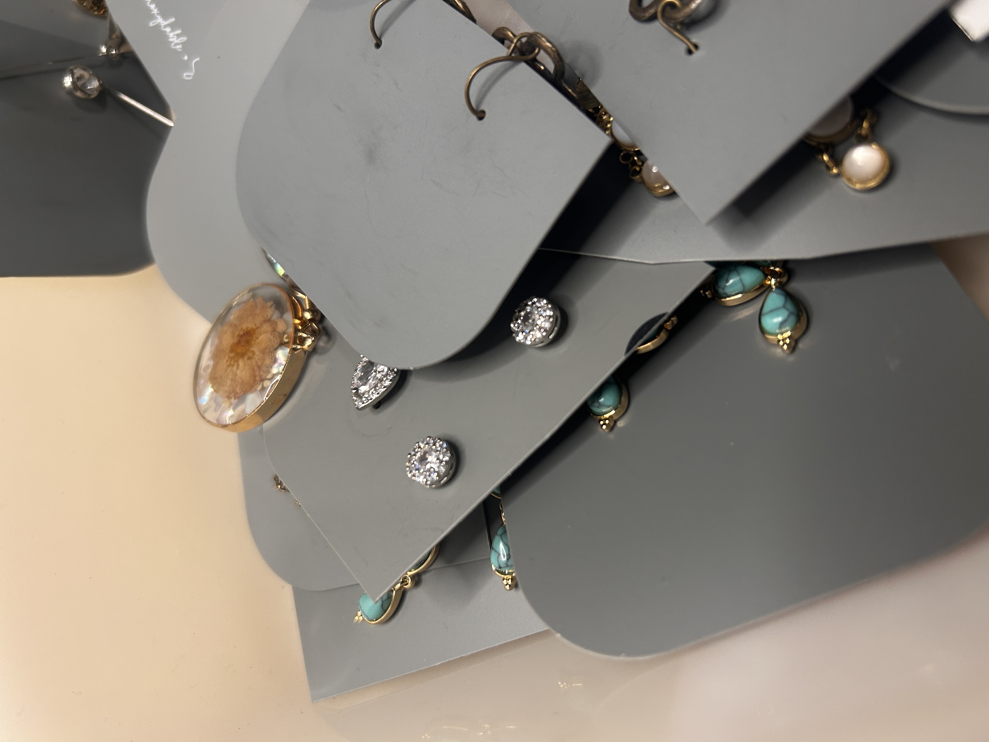 Toma detallada enfocada en pendientes de botón y pequeños pendientes colgantes, con acentos de cristal, una pieza única tipo ámbar y gotas de color turquesa en tarjetas grises.