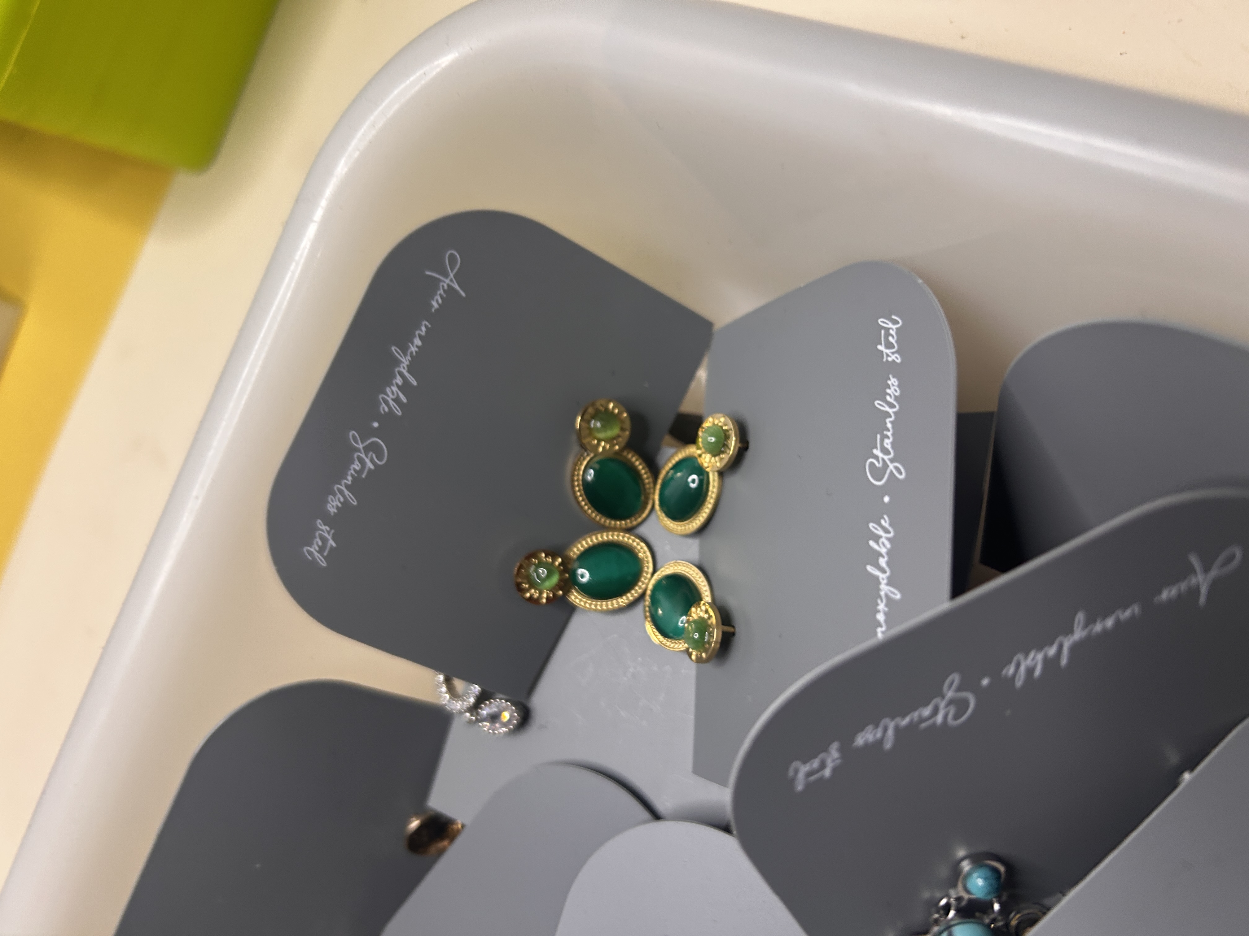 Primer plano de pendientes de acero inoxidable en tarjetas grises, destacando diseños de forma ovalada con piedras verdes vibrantes, algunos marcados como 'Acier inoxydable'.