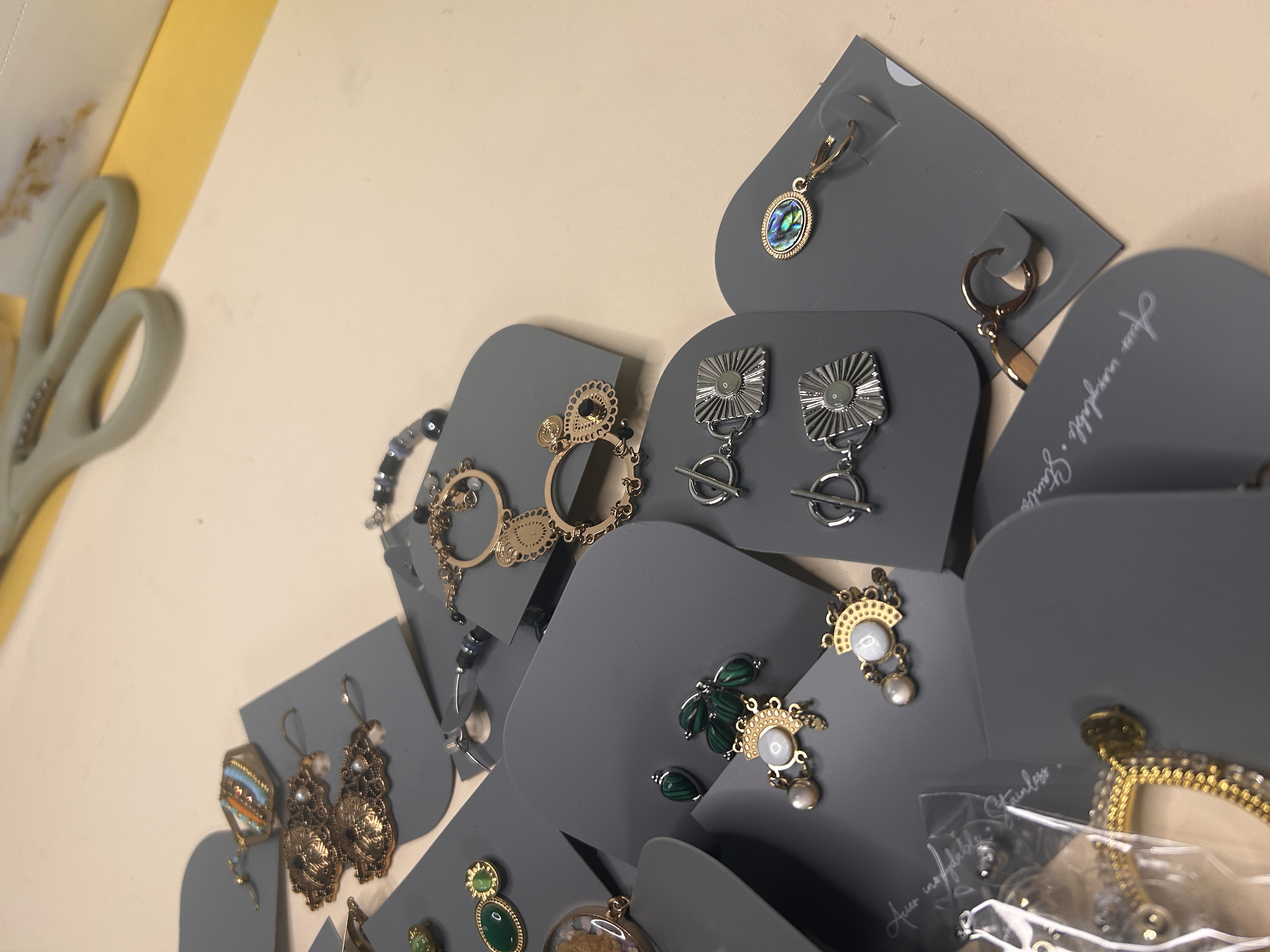 Toma de detalle de varios pares de pendientes de moda en tarjetas grises, mostrando diseños específicos que incluyen pendientes colgantes cuadrados, ovalados con piedras iridiscentes y elegantes detalles de perlas.