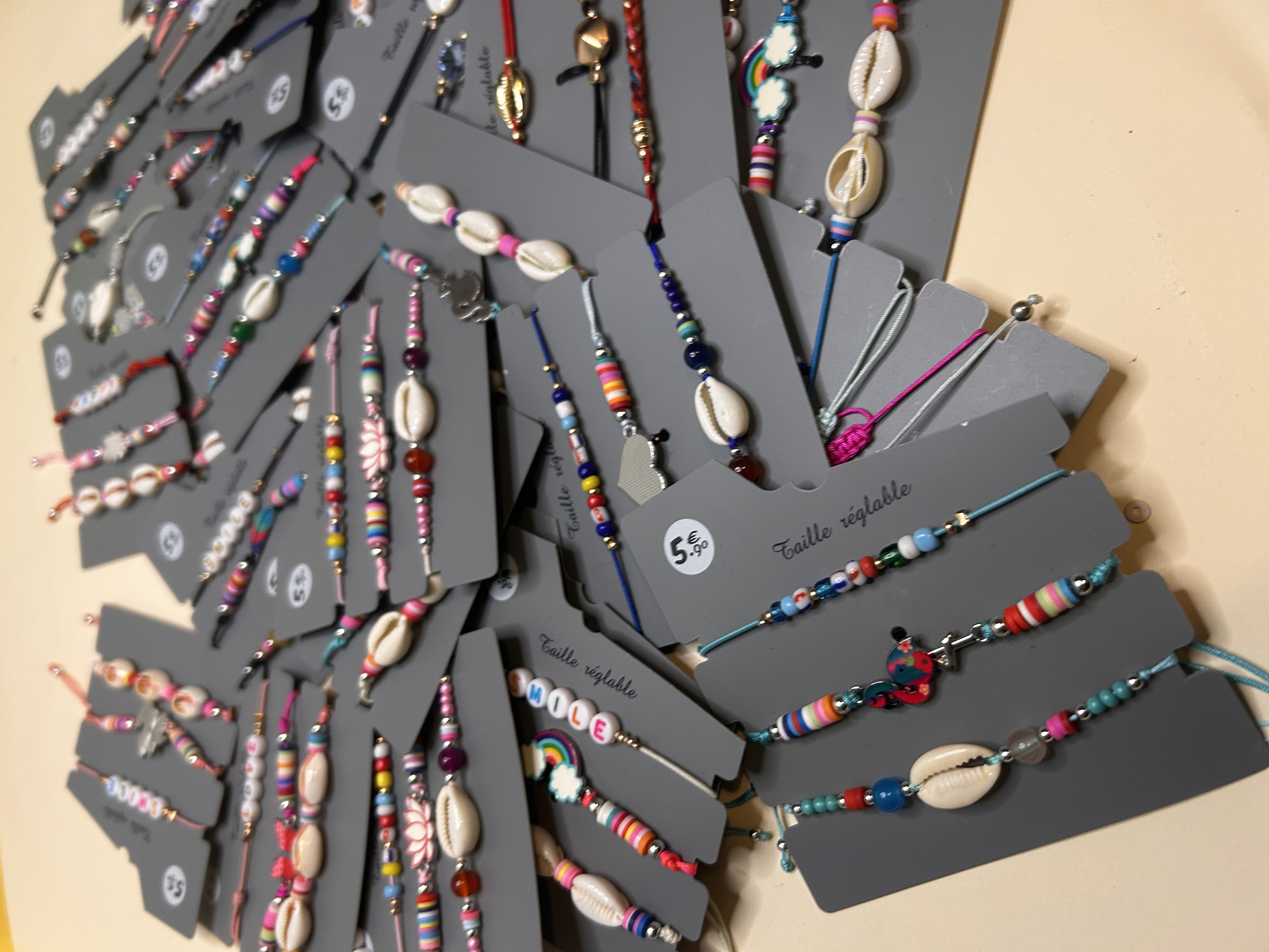 Vista angular de cerca de un montón de pulseras ajustables vibrantes, destacando el trabajo de abalorios, las conchas cowrie y algunas con abalorios 'SMILE', todas en cartones de presentación grises.