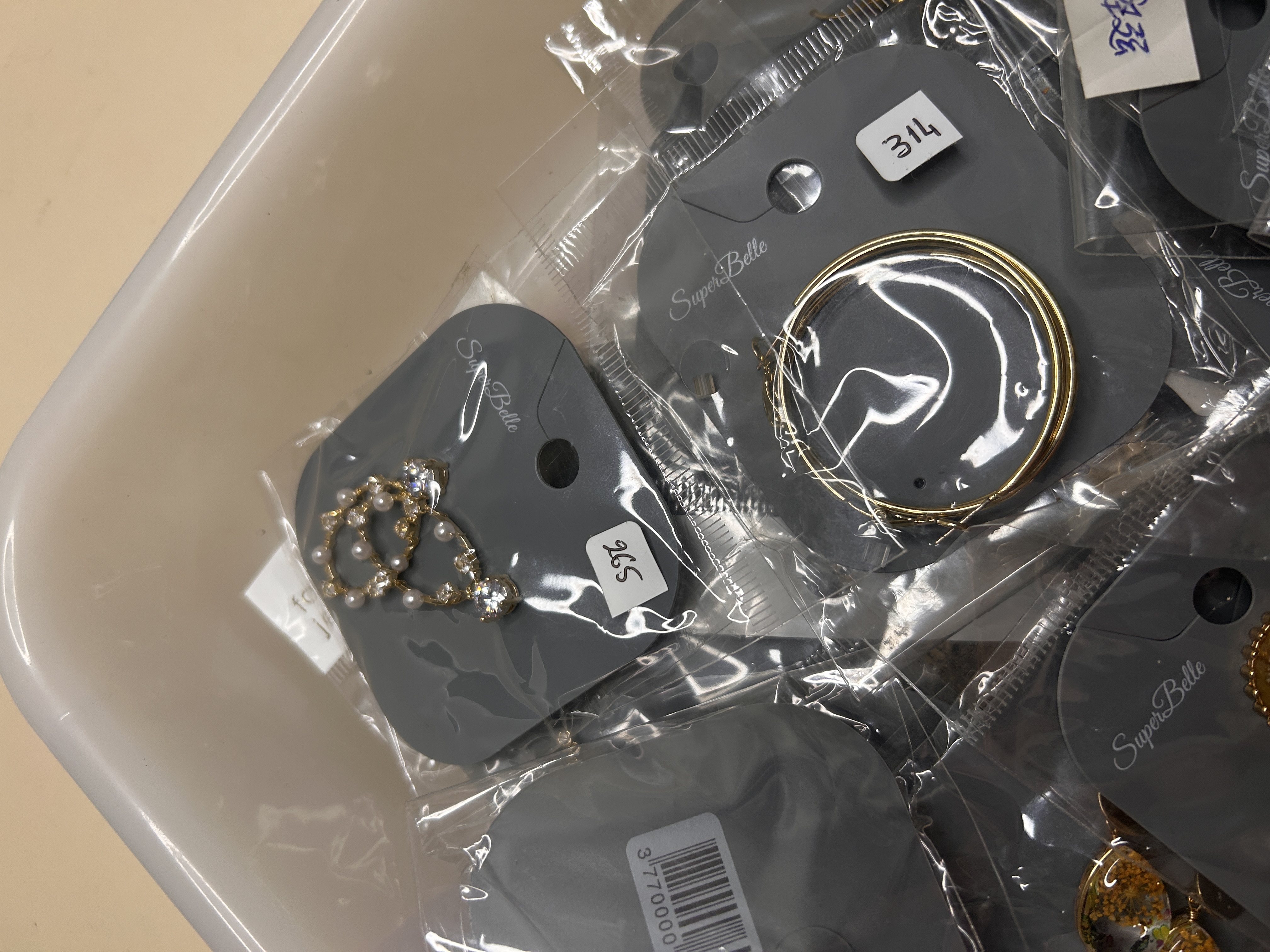 Primer plano que muestra un par de pendientes de aro en tono dorado y otro par de delicados pendientes colgantes con pequeñas perlas y cristales, todos sellados en plástico.