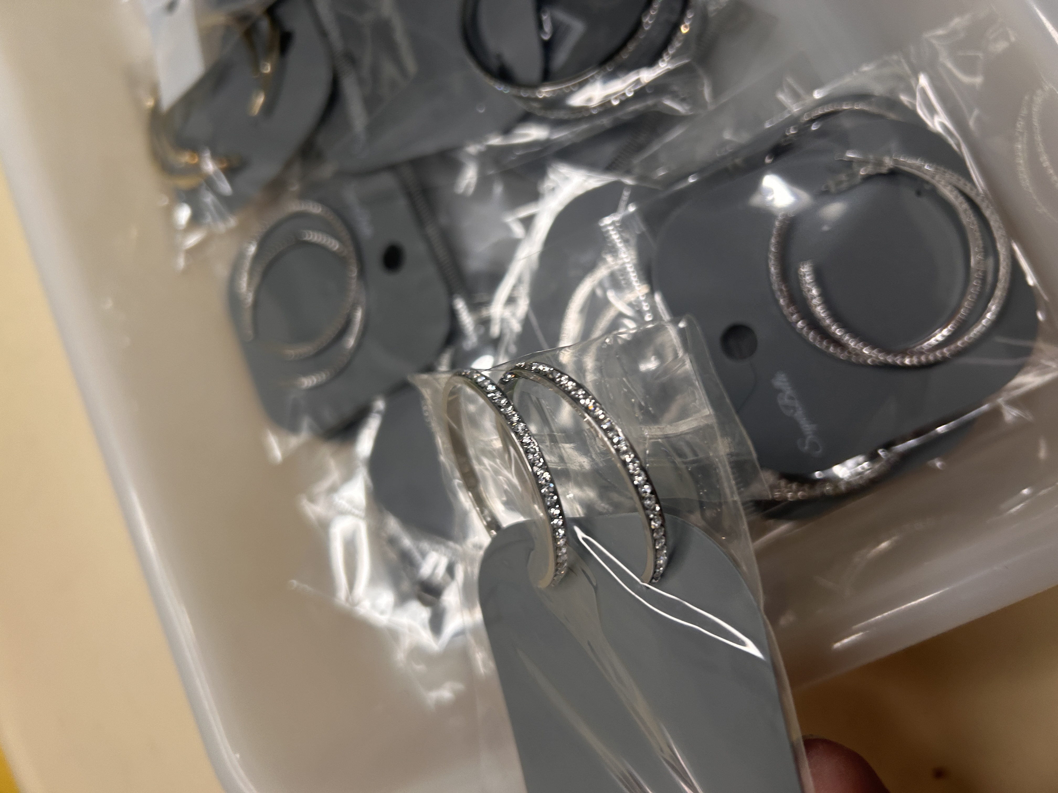 Mano sosteniendo un par de pendientes de aro plateados con detalles de cristales pavé, mostrando el embalaje individual dentro del lote BO-101.