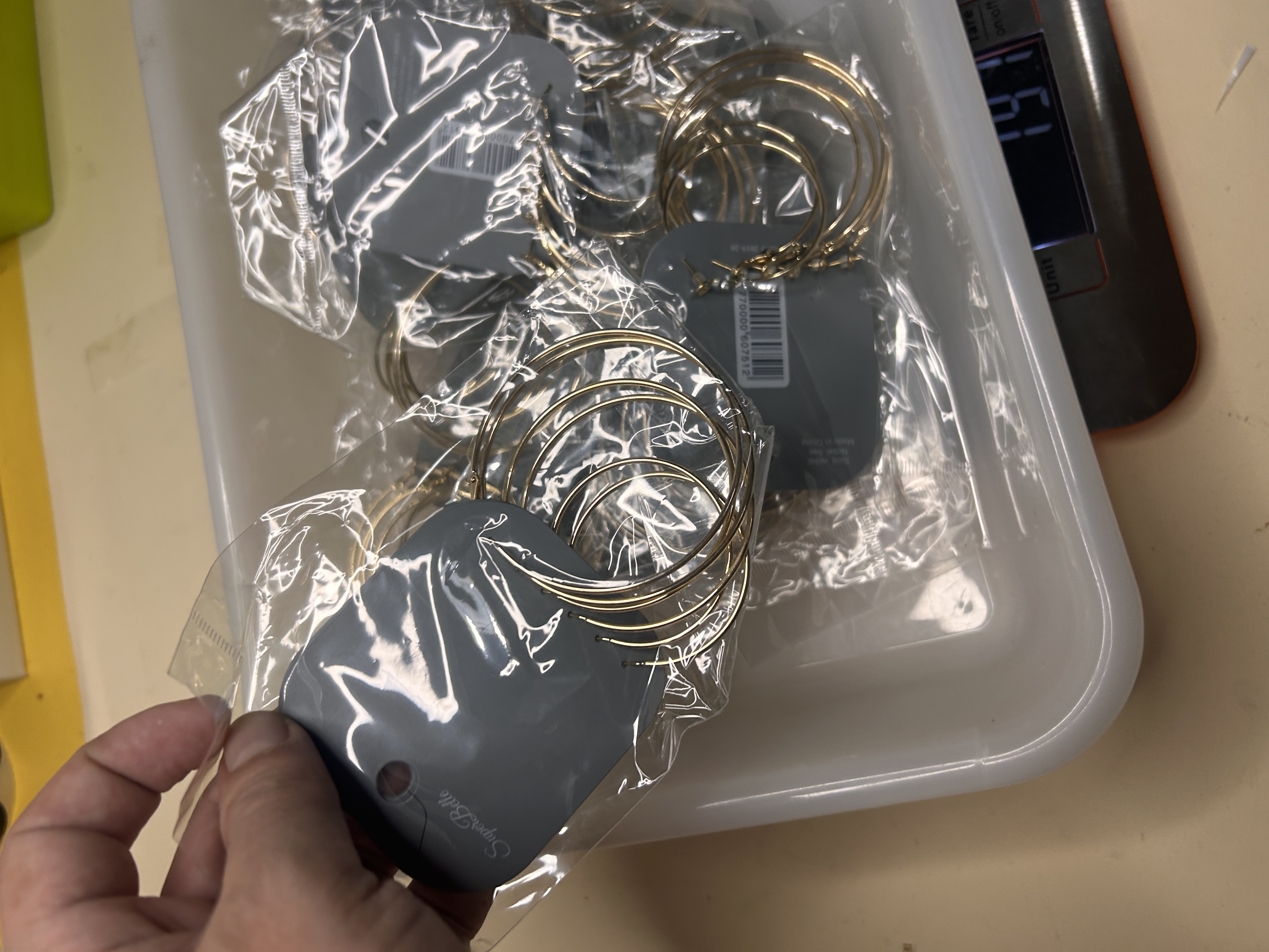 Primer plano de una mano cogiendo un set de pendientes de aro dorados empaquetado individualmente de la bandeja, con la báscula aún visible.
