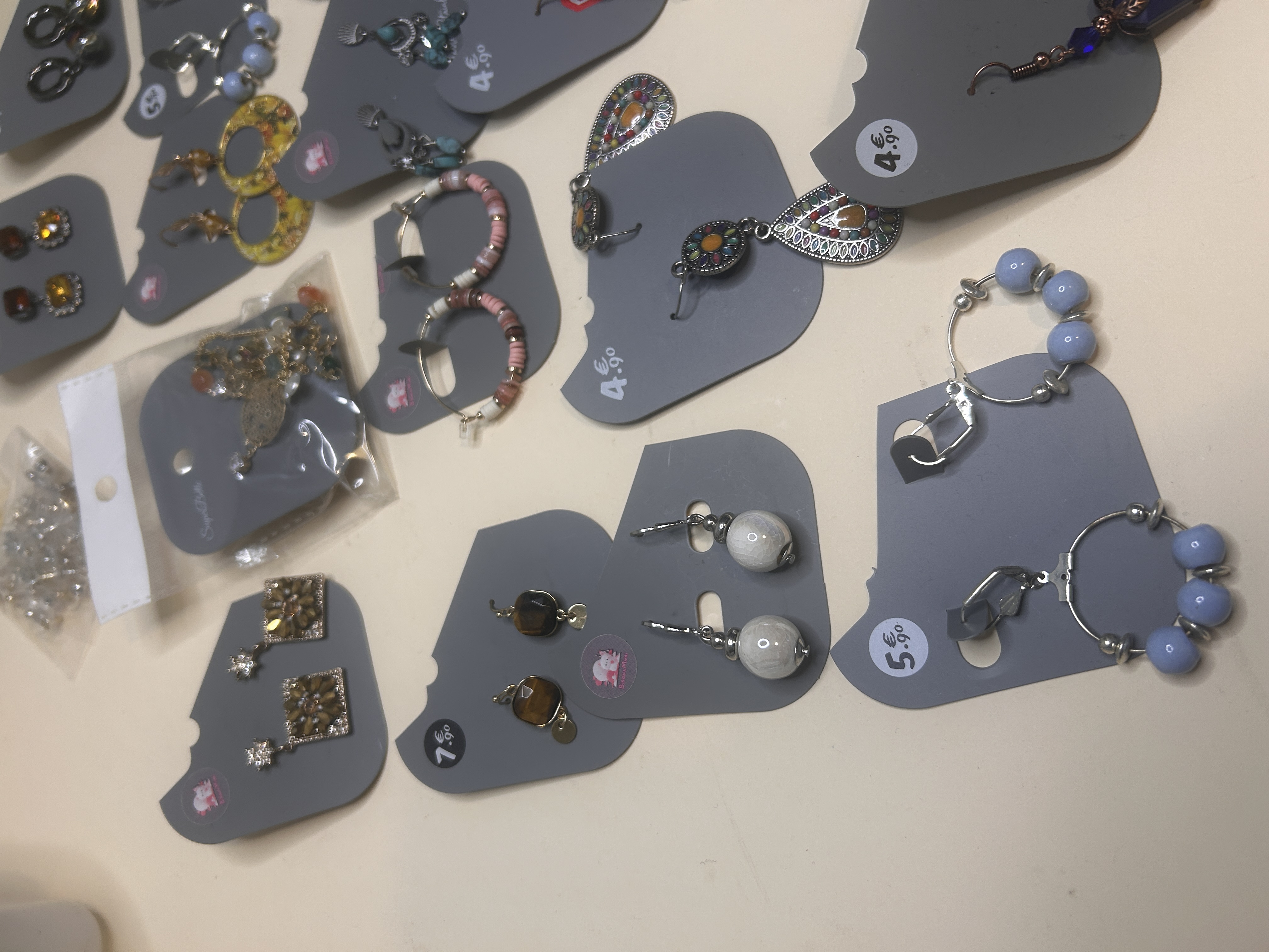 Toma de cerca de múltiples pendientes de bisutería en cartones grises, incluyendo aros con cuentas, colgantes con piedras y topes cuadrados, distribuidos sobre una superficie clara.