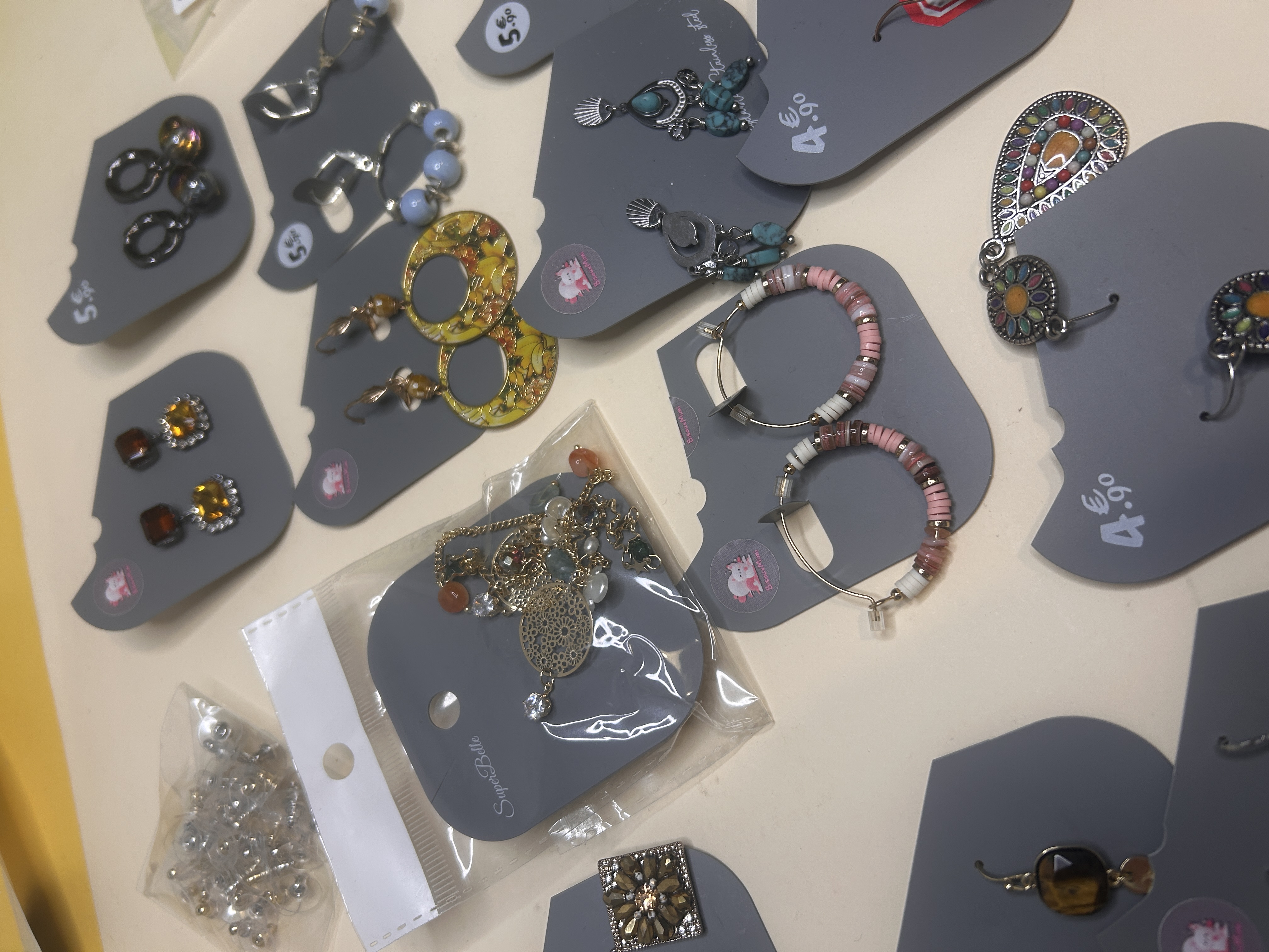 Una vista de cerca que captura una variedad de pendientes de bisutería en cartones de exhibición grises, destacando detalles como materiales mixtos, cuentas y diferentes formas. Algunas piezas están en bolsas de plástico transparente.