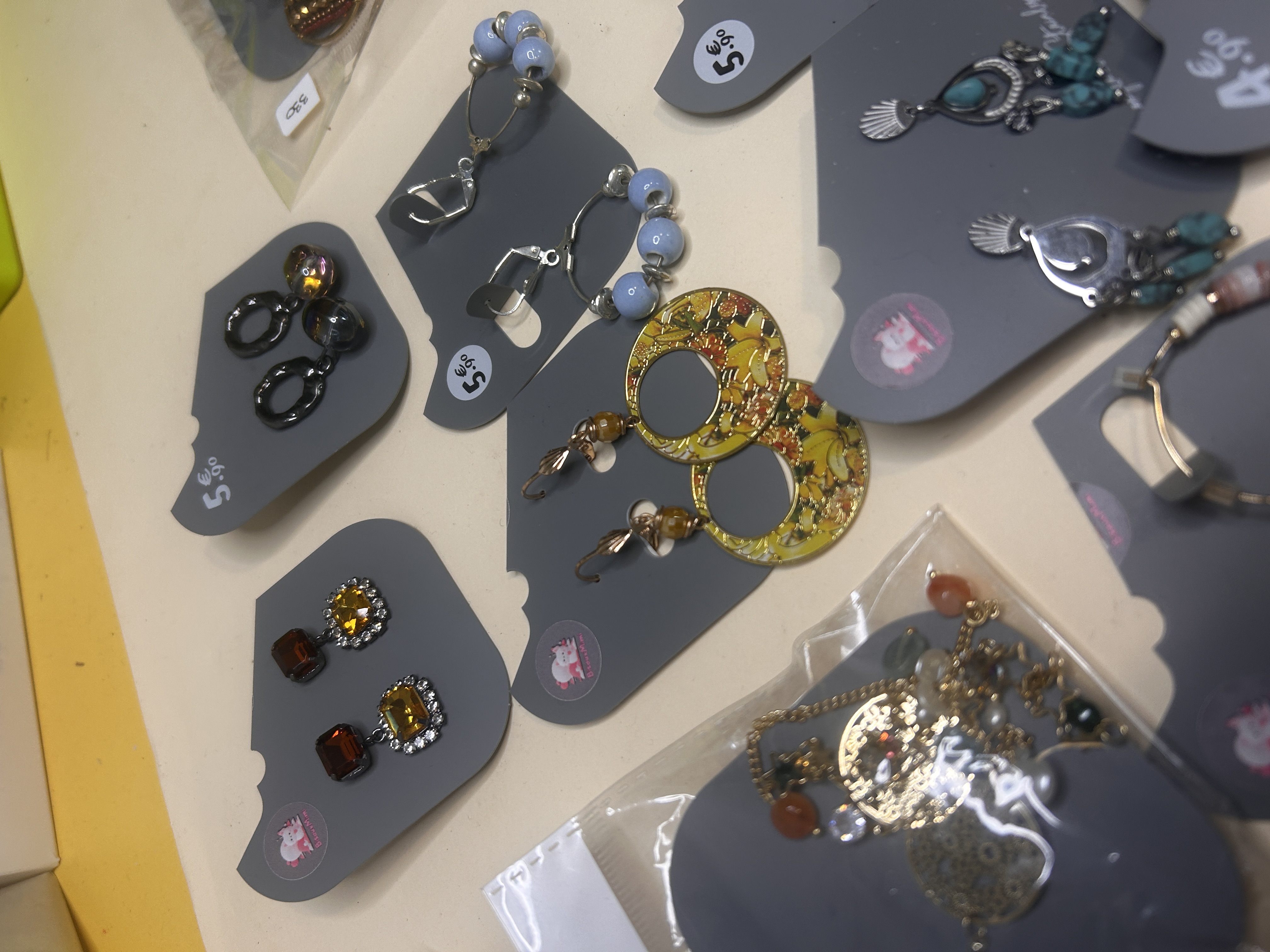 Toma detallada de varios pendientes de bisutería, incluyendo aros con estampado amarillo, pendientes con cuentas azules y topes en tonos ámbar, todos en cartones grises listos para la venta.