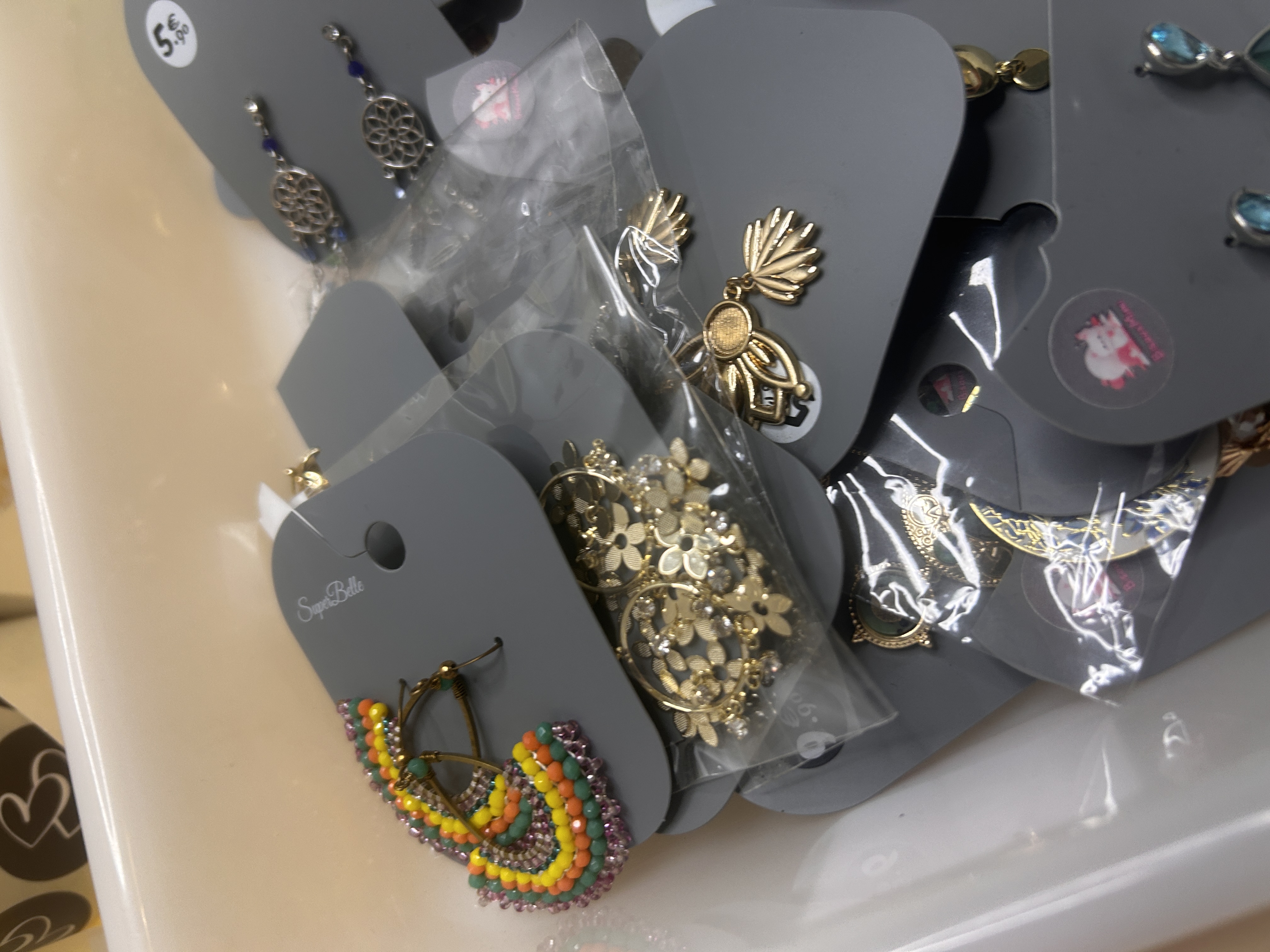Primer plano angular de pendientes de bisutería en una bandeja, mostrando un pendiente colorido en forma de C con cuentas y varios diseños florales y en forma de concha en tono dorado, todos empaquetados individualmente.