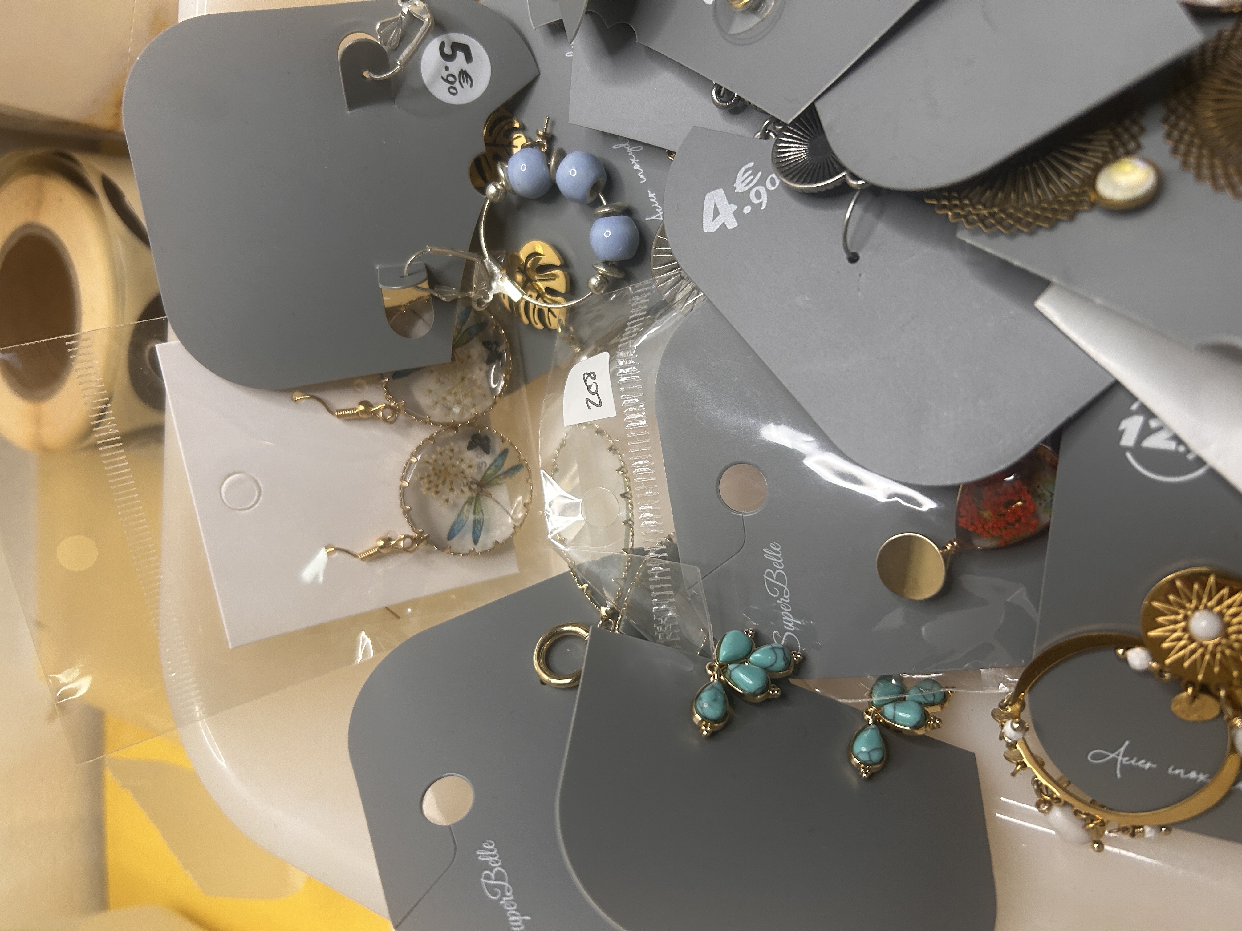 Toma detallada de pendientes en una bandeja, destacando pendientes colgantes de resina transparente con flores incrustadas, aros de cuentas azul claro y pequeños pendientes de tuerca con forma de abeja turquesa.