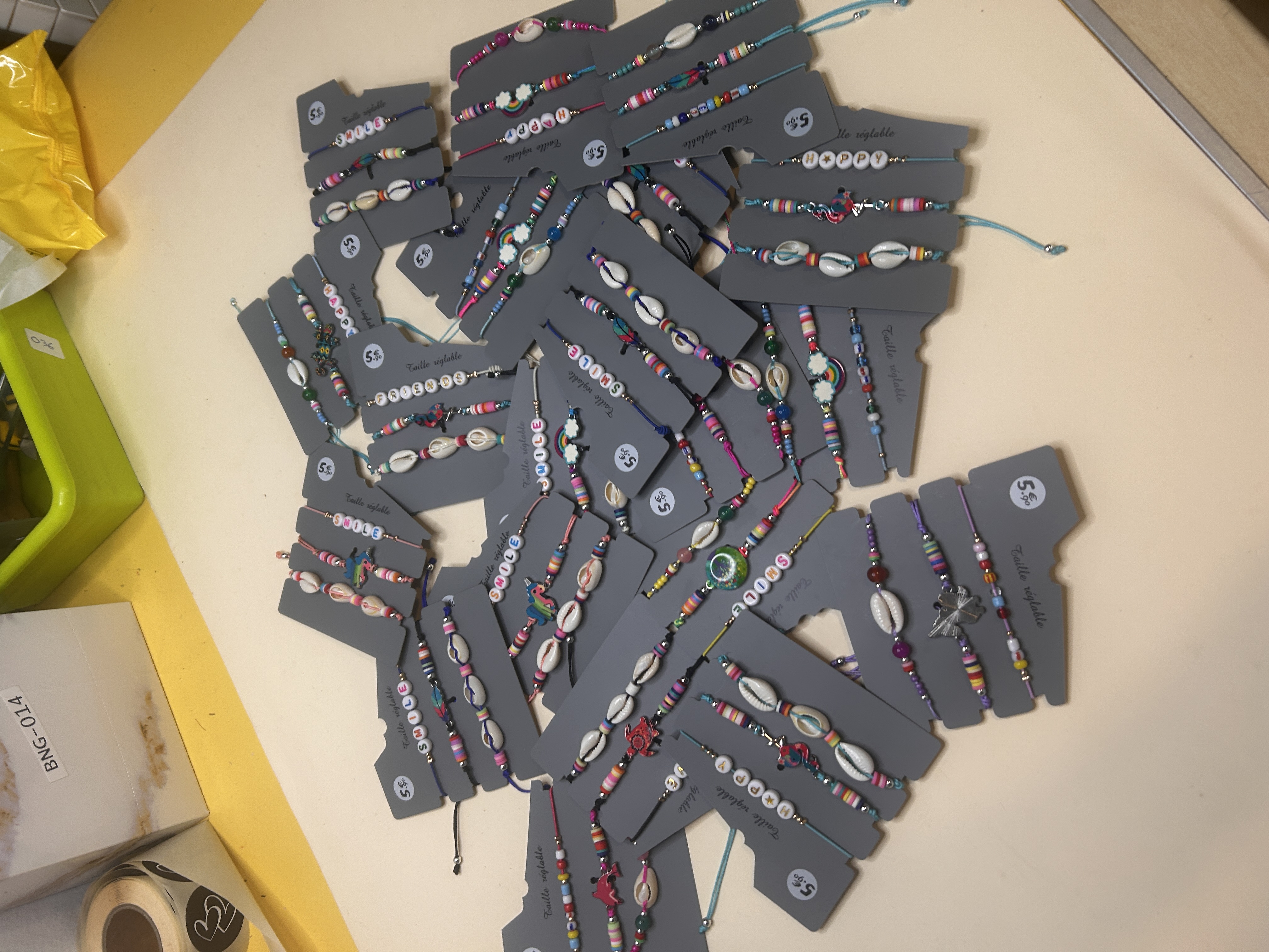 Una vista aérea que muestra una gran variedad de pulseras de bisutería, empaquetadas individualmente en tarjetas grises, extendidas sobre una superficie clara, con varios diseños de conchas y cuentas.