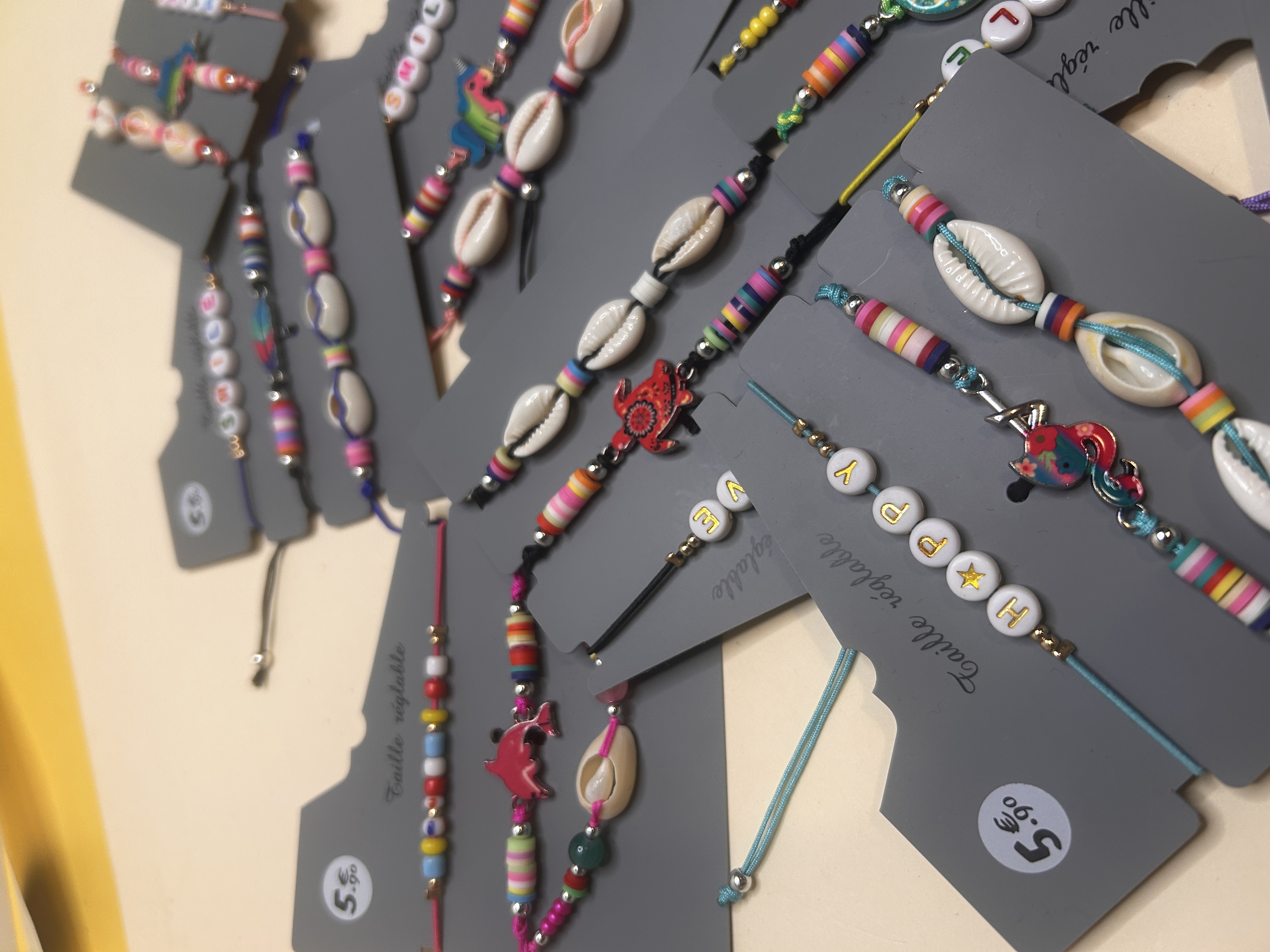 Otro primer plano de pulseras de bisutería en tarjetas, destacando diferentes diseños que incluyen un delfín rosa, una tortuga roja y cuentas con letras, todos con cordones y cuentas coloridos.