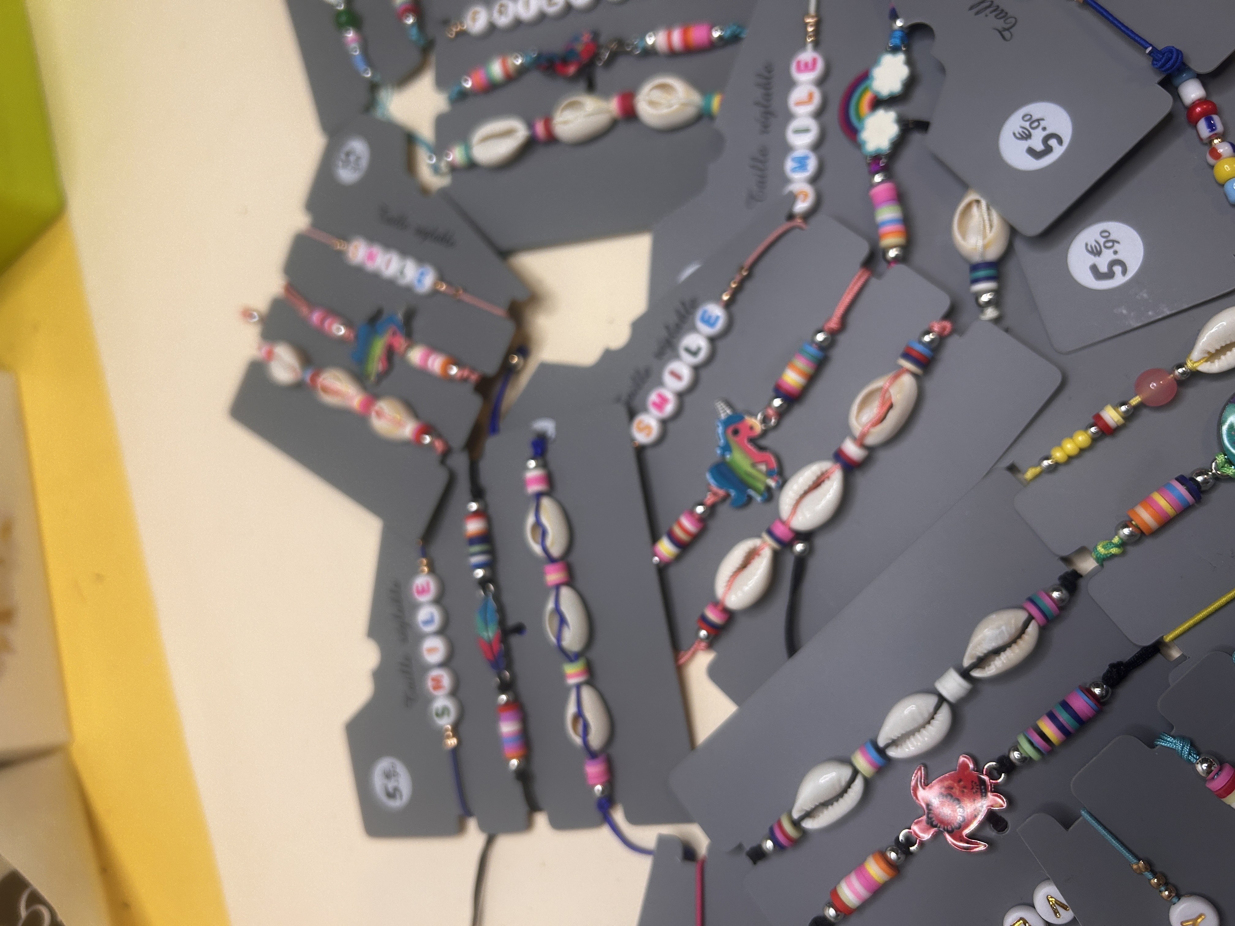 Una imagen de cerca que se centra en varias pulseras de bisutería coloridas, algunas con conchas de cauri, abalorios de arcoíris y cuentas con letras que forman 'DULCE', todas presentadas en tarjetas de exposición grises.